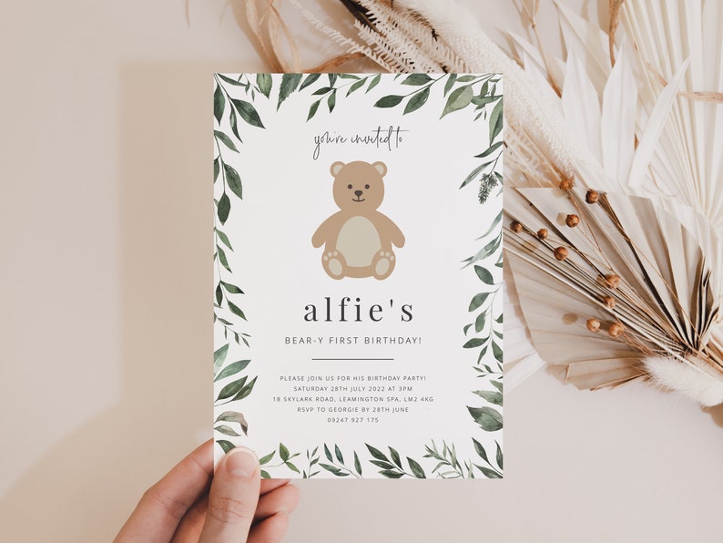 Puede incluir: Una tarjeta de invitaci&oacute;n blanca con un borde de hojas verdes. La tarjeta presenta un oso de peluche marr&oacute;n de dibujos animados y el texto "you're invited to alfie's BEAR-Y FIRST BIRTHDAY!" La invitaci&oacute;n incluye detalles para una fiesta de cumplea&ntilde;os, incluida la fecha, la hora, la ubicaci&oacute;n y la informaci&oacute;n de RSVP.