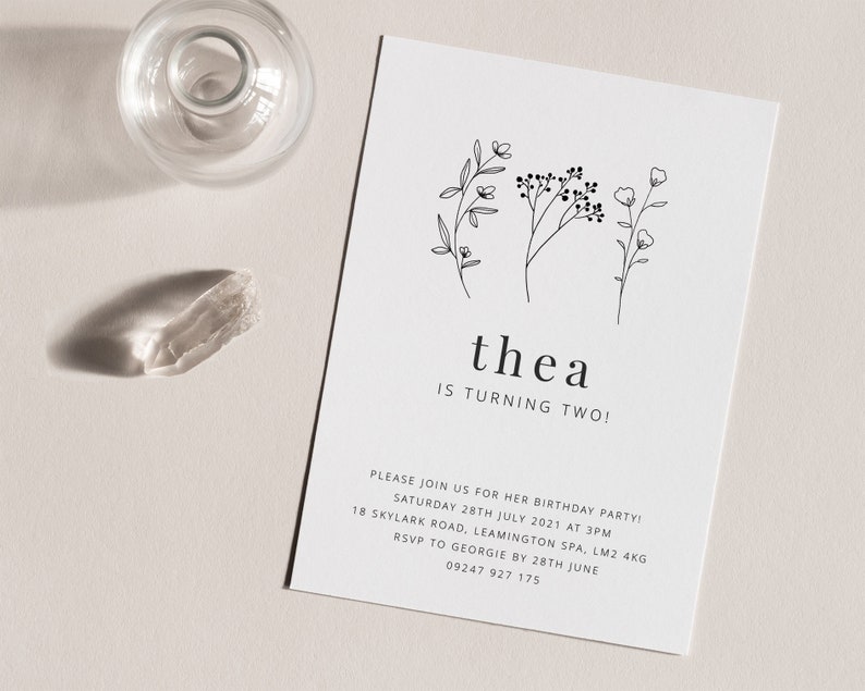 May include: A white card with black text and simple line drawings of flowers. The text reads "thea is turning two!" and includes details for a birthday party.