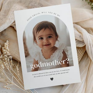 Könnte beinhalten: Eine weiße Karte mit dem Text "DEAR EMILIA, WILL YOU BE MY godmother?" zeigt ein lächelndes Baby-Mädchen. Die Karte enthält den Text "we feel so blessed to have you in our lives. It would mean the world to us if you would take on this special role!"
