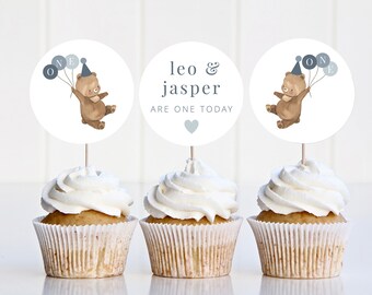 May include: Three white cupcakes with white frosting topped with round paper circles. The circles feature a brown bear wearing a blue party hat and holding blue balloons that say "ONE". The middle circle says "leo & jasper are one today" with a grey heart.