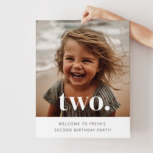 May include: A white poster with a photo of a young girl smiling at the beach. The text "two." is in large white letters on the poster. Below the photo, the text "Welcome to Freya's Second Birthday Party!" is in smaller white letters.
