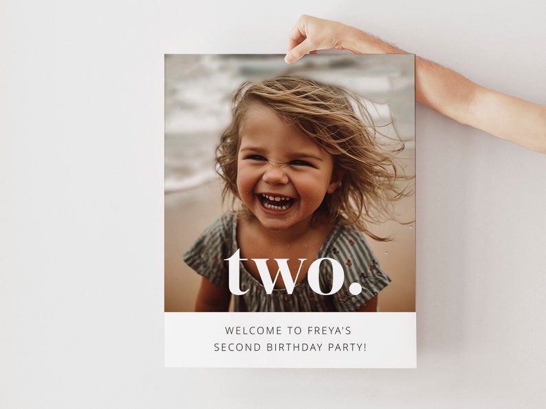 May include: A white poster with a photo of a young girl smiling at the beach. The text "two." is in large white letters on the poster. Below the photo, the text "Welcome to Freya's Second Birthday Party!" is in smaller white letters.