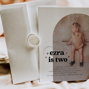 May include: A beige invitation card with a photo of a young boy sitting on a rock. The text on the card reads "CELEBRATE! +ezra+ +is two+" and includes details for a party.