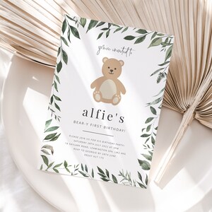 Puede incluir: Una tarjeta de invitaci&oacute;n blanca con un borde de follaje verde y una ilustraci&oacute;n de un oso de peluche marr&oacute;n. El texto dice "you're invited to alfie's BEAR-Y FIRST BIRTHDAY!" con detalles para la fiesta de cumplea&ntilde;os.