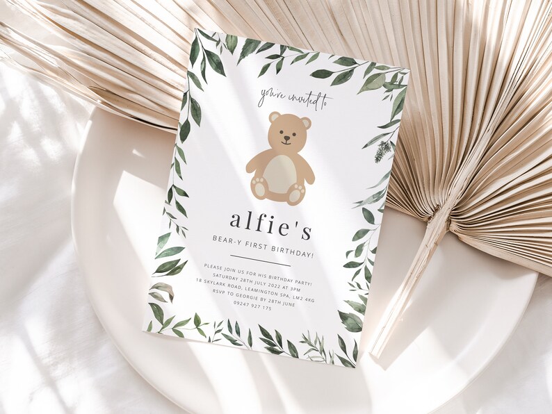 Puede incluir: Una tarjeta de invitaci&oacute;n blanca con un borde de follaje verde y una ilustraci&oacute;n de un oso de peluche marr&oacute;n. El texto dice "you're invited to alfie's BEAR-Y FIRST BIRTHDAY!" con detalles para la fiesta de cumplea&ntilde;os.