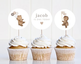 May include: Three brown bear themed cupcake toppers with white frosting. The toppers feature a bear holding balloons that say "ONE", a bear holding a flag, and a topper that says "jacob is one today" with a small pink heart.