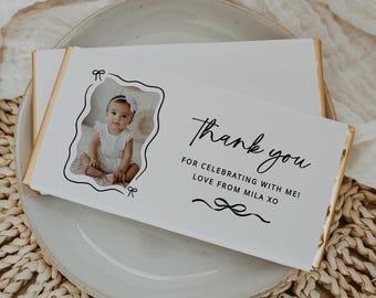 May include: White chocolate bar with a gold foil wrapper. The bar has a photo of a baby girl in a white dress and the text "Thank you for celebrating with me! Love from Mila XO".