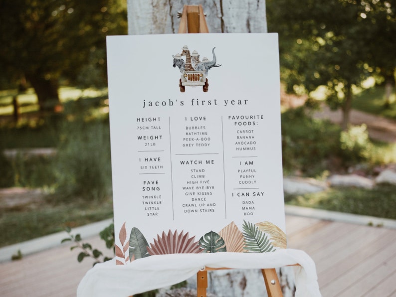 May include: A white sign with a jungle theme, featuring a cartoon jeep with animals. The sign lists a child's first year milestones, including height, weight, favorite foods, songs, and things they can do. The text reads "jacob's first year".