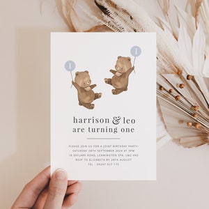 May include: A white invitation card with a blue and brown illustration of two bears holding balloons with the number "1" on them. The text on the card reads "harrison & leo are turning one" and includes details for a joint birthday party.