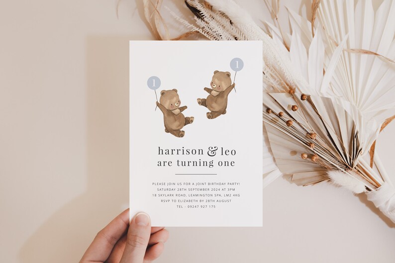 May include: A white invitation card with a blue and brown illustration of two bears holding balloons with the number "1" on them. The text on the card reads "harrison & leo are turning one" and includes details for a joint birthday party.