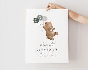 May include: A white poster with a brown bear holding balloons that spell out "ONE". The text "welcome to greyson's bear-y first birthday party!" is printed below the bear.