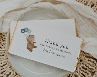 May include: A white chocolate bar with gold foil wrapping. The bar has a thank you message for coming to a party, with the text "thank you for coming to my party! love from oscar xo". The bar also features a cartoon illustration of a brown bear holding blue balloons with the letters "ONE" on them.