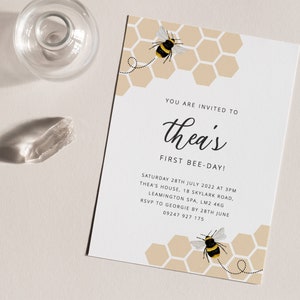 K&ouml;nnte beinhalten: Eine Einladung zu einer ersten Geburtstagsfeier zum Thema Bienen mit einem Wabenmuster und zwei Comic-Bienen. Der Text der Einladung lautet "You are invited to Thea's First Bee-Day!" mit Datum, Uhrzeit und Ortsangaben darunter.