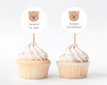 May include: Two white cupcakes with brown bear toppers. One topper says "jasper is one" and the other says "happy birthday!"