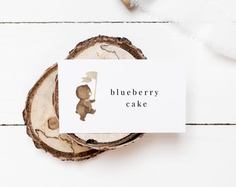 May include: A white card with the words "blueberry cake" printed in black. A watercolor illustration of a brown bear holding a flag is on the card. The card rests on a wooden slice with a white background.
