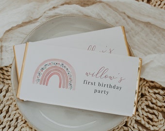 Könnte beinhalten: Zwei weiße Schokoriegelverpackungen mit goldenen Rändern sind auf einem grauen Teller gestapelt. Die obere Verpackung zeigt ein Aquarell-Regenbogen-Design und den Text "willow's first birthday party" in rosa Schrift.
