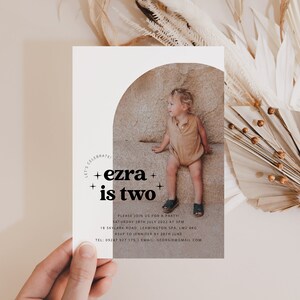 May include: A white invitation card with a photo of a young child in a tan outfit sitting on a rock. The text on the card reads "Let's Celebrate + ezra + is two +" and includes details for a birthday party.