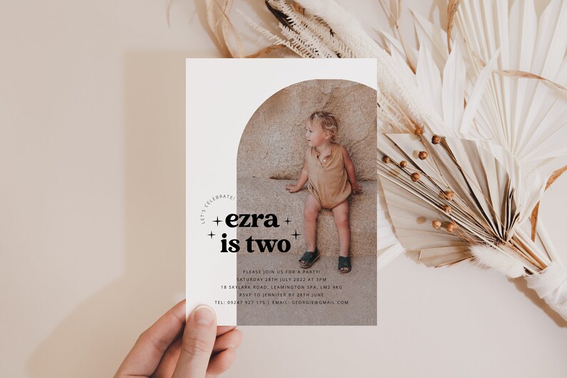 May include: A white invitation card with a photo of a young child in a tan outfit sitting on a rock. The text on the card reads "Let's Celebrate + ezra + is two +" and includes details for a birthday party.