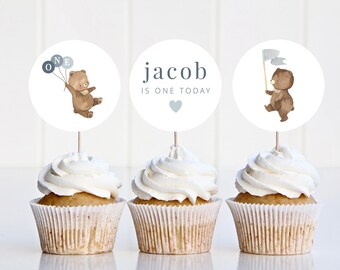 May include: Three cupcakes with white frosting and brown paper cases. Each cupcake has a round, white paper topper with a brown bear illustration. The toppers say "ONE", "jacob is one today", and a bear holding a flag.