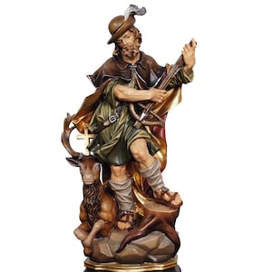 May include: A carved wooden statue of a man in a hat and cape, holding a crossbow. The statue is brown, green, and gold, with a deer at the man's feet. The man is standing on a tree stump.