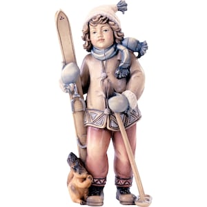 May include: A wooden figurine of a young skier in winter attire. The figure wears a tan coat, pink pants, a white hat, and a blue scarf. The skier holds skis and poles, with a small squirrel at their feet.