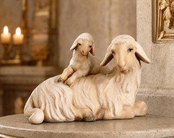 May include: A hand-carved wooden figurine depicting a mother sheep lying down with a small lamb resting on its back. The sculpture features natural light wood tones with detailed textures representing the wool, ears, and facial features of both animals.
