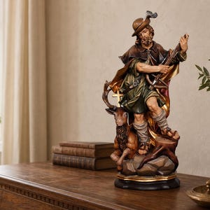 May include: A hand-painted figurine of Saint Hubertus wearing a brown hat with a feather, a green tunic, and a brown cloak. The figure holds a crossbow and stands beside a stag with a golden cross between its antlers, all set on a dark base.