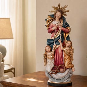 May include: A multi-colored statue of the Virgin Mary untying knots in a rope, wearing a red dress and blue cloak with a gold halo. Two winged cherubs stand at the base near a coiled snake, all resting upon a crescent moon and cloud base.