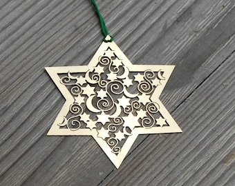 May include: A gold-coloured, six-pointed star ornament with a cut-out design of stars, moons, and swirls. The ornament is hanging from a green string.