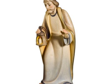 May include: A wooden figurine of a man in a long robe, holding a lantern with a lit candle. The figure has a beard and is looking down. The robe is cream-colored with gold trim, and the lantern is brown. A religious-themed item.