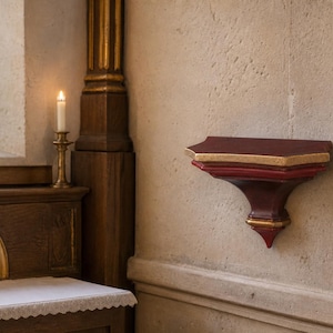 May include: A wall-mounted wooden shelf featuring a deep red finish with gold-painted edges. The shelf has a decorative, tapered bracket design and is attached to a textured, light-colored stone wall near a wooden structure with a lit candle.