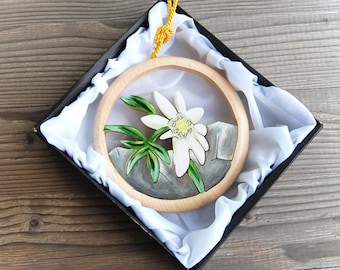May include: A wooden round ornament featuring a white edelweiss flower and green leaves against a grey background. The ornament is presented in a black box with white tissue paper.