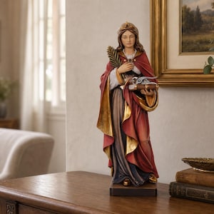 Può includere: Una statuetta in piedi raffigurante una donna con un mantello rosso e oro sopra un abito scuro, che tiene una foglia di palma in una mano e un libro con sopra un attrezzo metallico nell'altra. La figura indossa una corona ed è su una base.