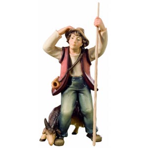 May include: A wooden figurine of a shepherd with a red vest, blue pants, and a brown hat. He is holding a staff and looking up. A brown and white goat is sitting behind him.