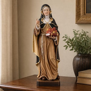 Op de afbeelding: Een staand religieus beeldje van een non in een zwart habijt over een beige gewaad. De figuur houdt een rood boek met een boeket van rode en oranje bloemen in de arm, terwijl de andere hand is opgeheven met de wijsvinger naar boven gericht.