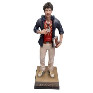 May include: A figurine of Carlo Acutis, a young man wearing a blue jacket, red shirt with a gold heart, and white pants. He is holding a book in his right hand and has a gold chain around his neck. The figurine is standing on a base with the words "CARLO ACUTIS" written on it.