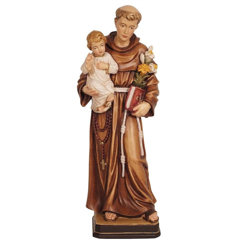 May include: A wooden statue of Saint Anthony of Padua holding a baby Jesus in his arms. The statue is painted in shades of brown and white, and the baby Jesus is wearing a white robe. Saint Anthony is holding a book in his left hand and a rosary in his right hand.