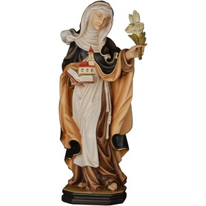Può includere: Una statua di legno di una santa, vestita con una tunica nera e marrone, che tiene un libro e un fiore bianco. La statua è in piedi su una base di legno.