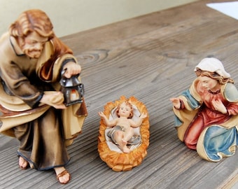May include: A nativity scene with a wooden figurine of Joseph holding a lantern, Mary kneeling and looking at the baby Jesus, and a baby Jesus figurine lying in a manger.