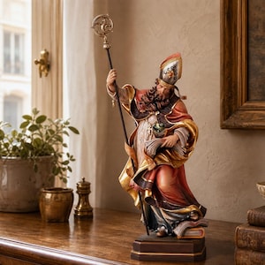 Könnte beinhalten: Eine handbemalte Bischofsfigur mit einer rot-goldenen Mitra und prunkvollen Gewändern, die einen hohen Stab mit dekorativem Aufsatz hält. Die Figur steht auf einem rechteckigen Sockel und zeigt detaillierte Gesichtszüge, einen Bart und Falten.