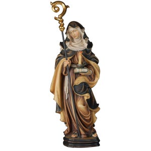 May include: A wooden statue of a female saint holding a book and a lit candle. She is wearing a brown and black robe and a white head covering. She is holding a staff with a golden crook.