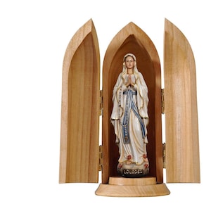 Könnte beinhalten: Ein Holzschrein mit zwei geöffneten Türen, der eine weiße Statue der Jungfrau Maria enthüllt, die auf einem Sockel mit der Aufschrift "Lourdes" steht.