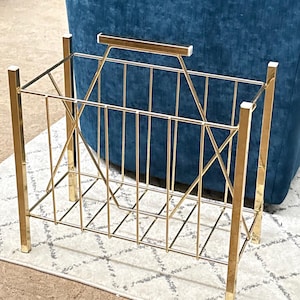 May include: A gold-colored metal magazine rack with a rectangular frame and a handle. The rack has a geometric design with vertical and diagonal bars. It is placed on a patterned rug, with a blue armchair in the background.