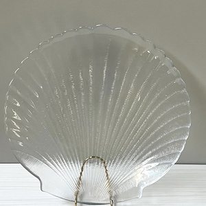 May include: A clear glass serving dish shaped like a seashell with a gold metal stand. The dish has a ribbed texture.