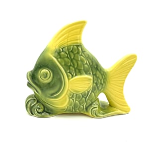 May include: A ceramic fish figurine in shades of green and yellow. The fish has a textured, scale-like pattern on its body, with a yellow dorsal fin and tail. The figurine is a decorative item, likely for home decor.