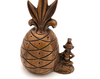 May include: A brown, carved pineapple figurine with a detailed design. A small figure sits beside the pineapple. The figurine has a textured surface and is set against a white background. The pineapple's top has a detailed leaf design.
