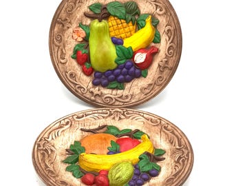 May include: Two decorative ceramic plates with raised relief fruit designs. Each plate features a variety of colorful fruits, including bananas, grapes, strawberries, and a pear, set against a brown, textured background with ornate edges.