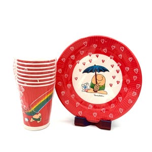 May include: A set of red and white Tom Wilson themed tableware. The set includes a stack of cups with a rainbow design and a plate with a cartoon character under an umbrella. The plate has a red border with heart accents.