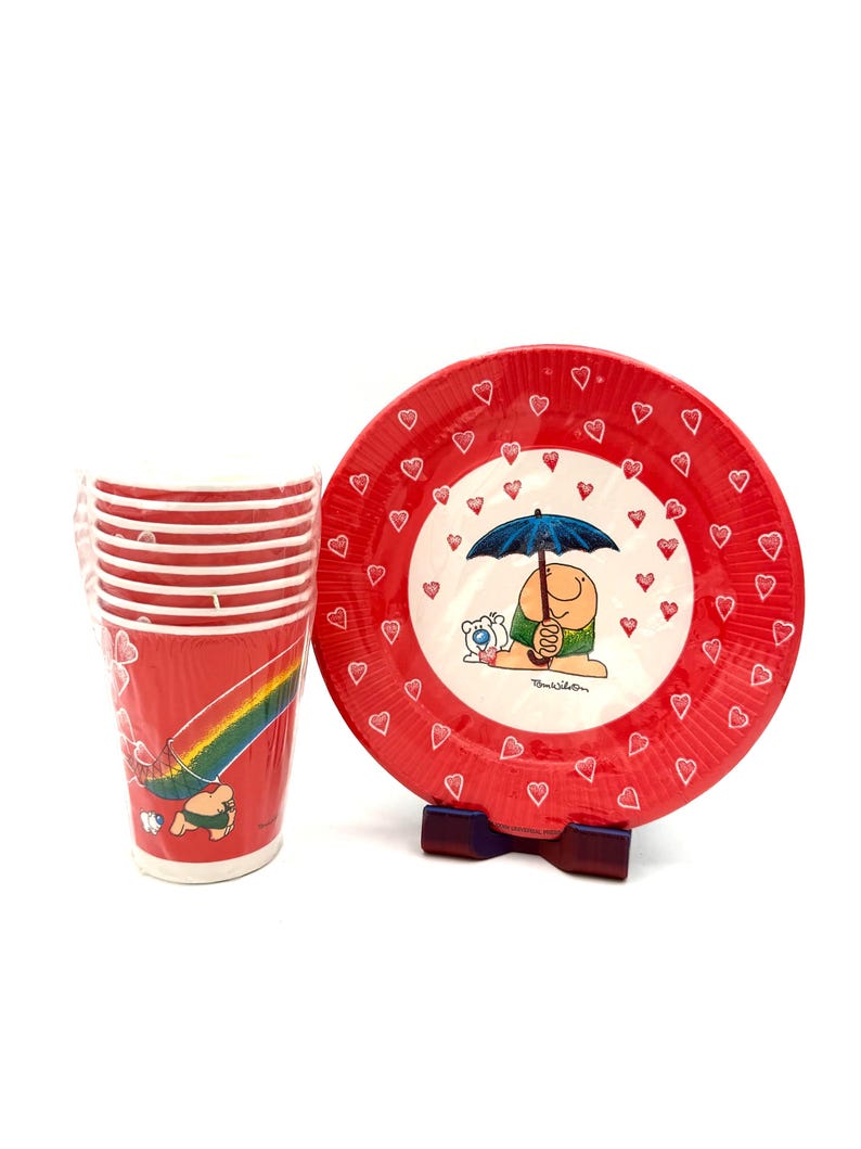 May include: A set of red and white Tom Wilson themed tableware. The set includes a stack of cups with a rainbow design and a plate with a cartoon character under an umbrella. The plate has a red border with heart accents.