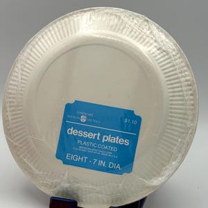 May include: A stack of eight white plastic-coated dessert plates, each 7 inches in diameter, wrapped in clear plastic. A blue label reads "dessert plates" and "plastic coated". The brand is Americard American Greetings.